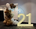 A 21st birthday party decoration setup featuring a large illuminated marquee '21' on the floor, a balloon arch in brown, gold, and white, and a circular backdrop with a white script sign reading 'Twenty One' in an indoor event space.